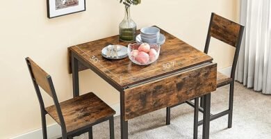 Juego de comedor compacto plegable de estilo industrial con mesa de madera extensible y dos sillas, decorado con un jarrón, platos y frutas, ubicado en un espacio iluminado.