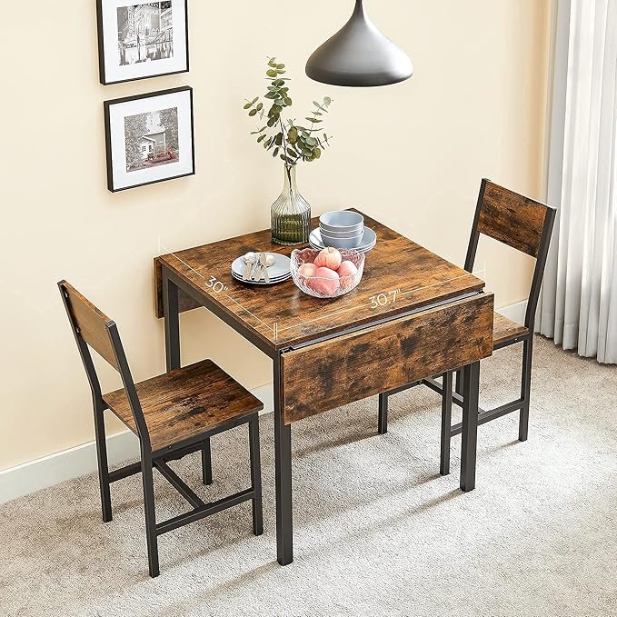 Juego de comedor compacto plegable de estilo industrial con mesa de madera extensible y dos sillas, decorado con un jarrón, platos y frutas, ubicado en un espacio iluminado.