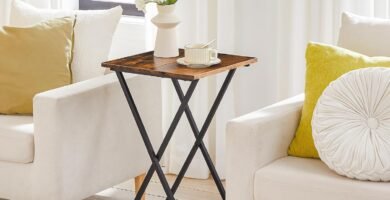 Mesa auxiliar plegable de madera con estructura metálica negra, decorada con un jarrón blanco y una taza, situada entre dos sillones blancos con cojines en tonos neutros y amarillos.