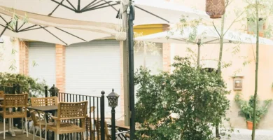 Terraza de cafetería al aire libre con mesas plegables y sillas de madera y mimbre, grandes sombrillas blancas y plantas decorativas en un patio empedrado.