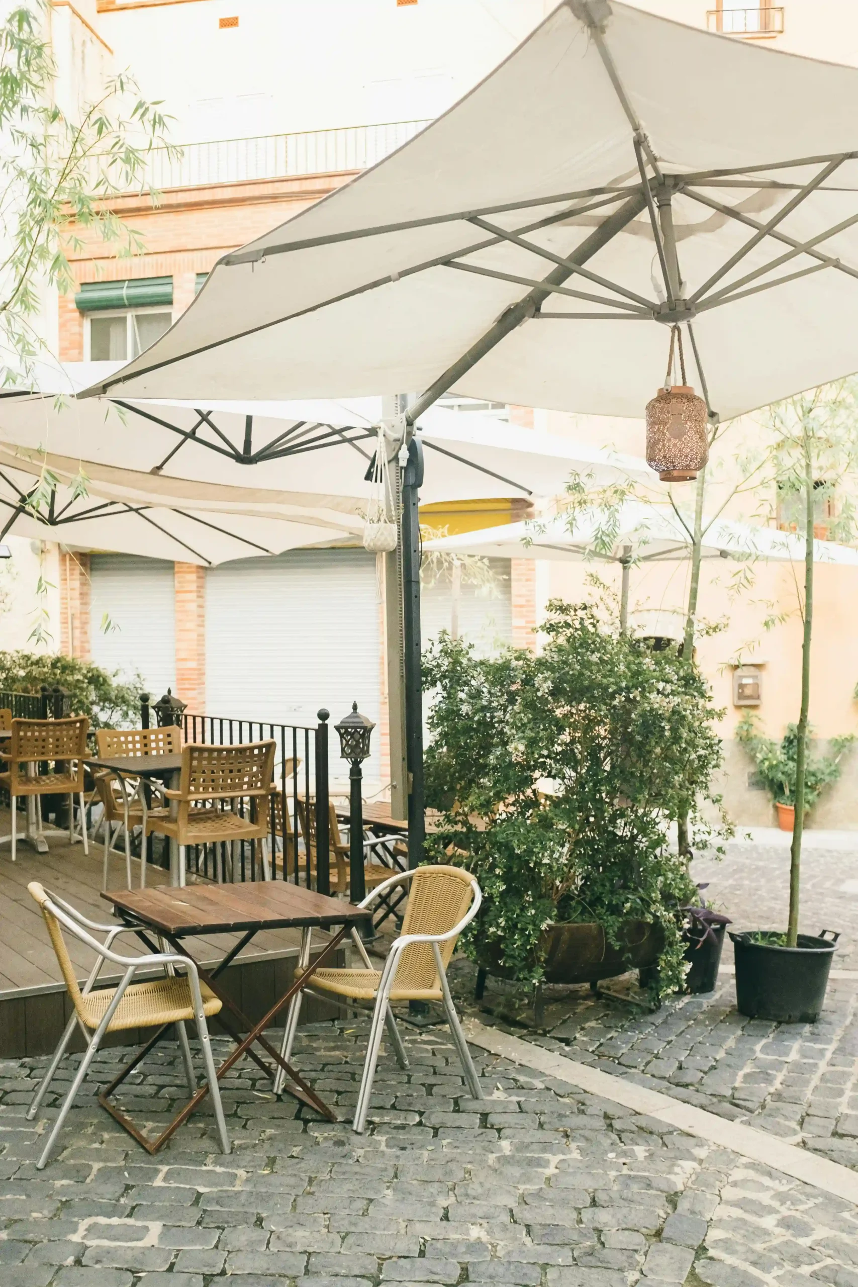 Terraza de cafetería al aire libre con mesas plegables y sillas de madera y mimbre, grandes sombrillas blancas y plantas decorativas en un patio empedrado.