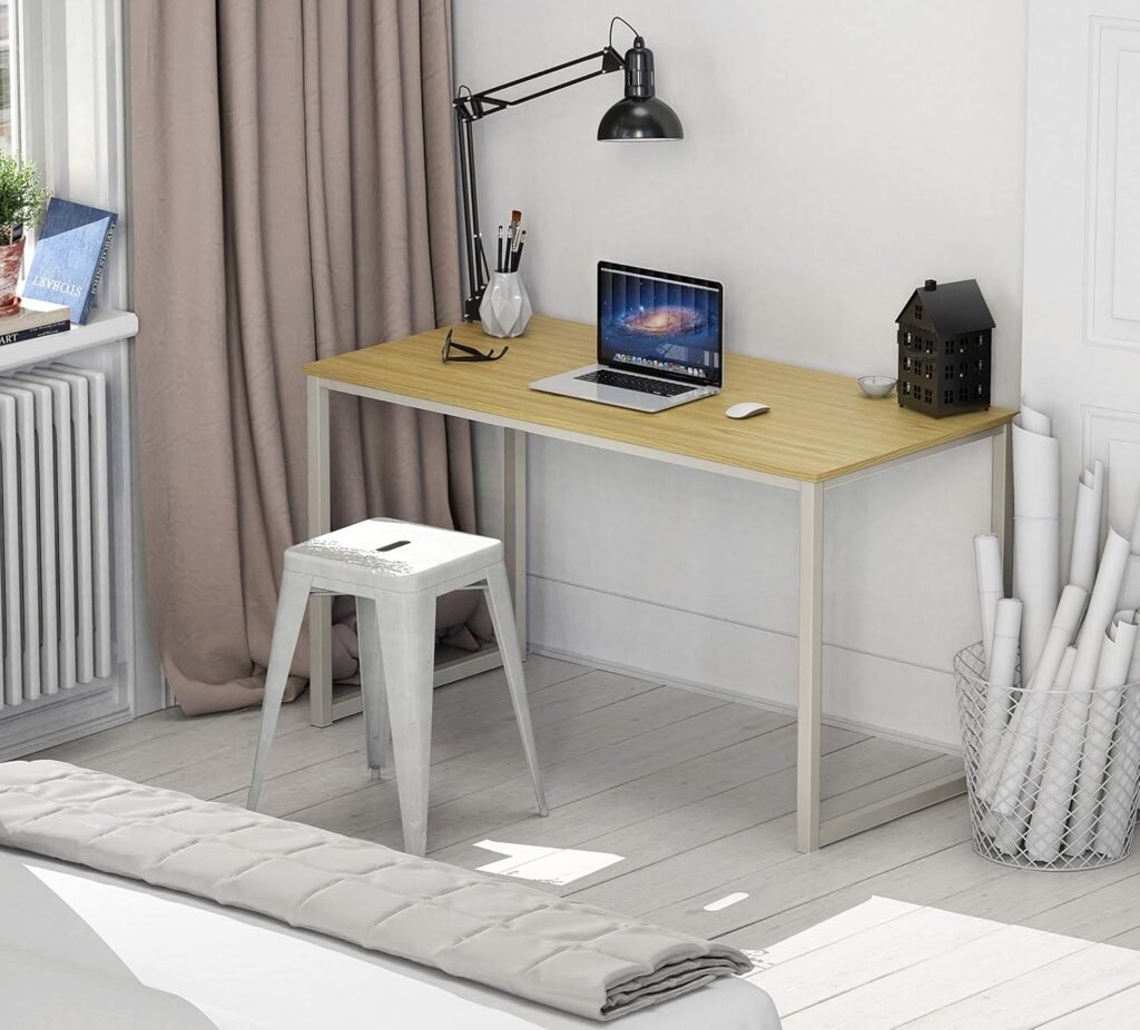 Escritorio moderno con superficie de madera clara y estructura metálica blanca, ubicado en una habitación con luz natural, equipado con laptop, lámpara de brazo, taburete metálico y objetos decorativos.