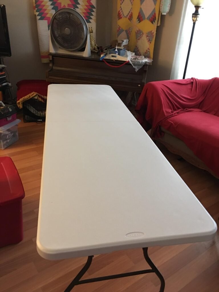 Mesa plegable blanca Cosco en una sala de estar, rodeada de varios objetos y muebles.