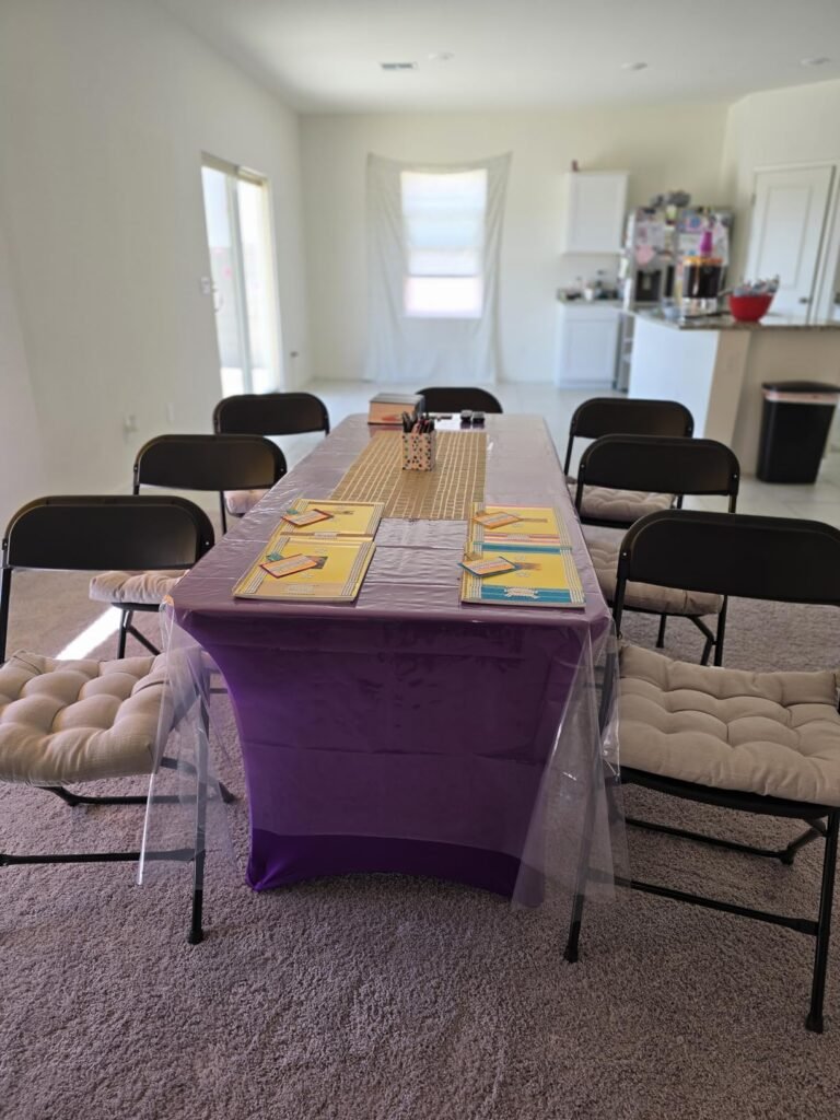Mesa de seis pies decorada para actividad de manualidades en un salón, con sillas y materiales de dibujo.