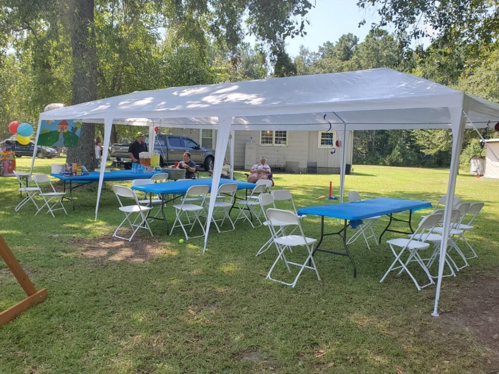Fiesta al aire libre con mesas plegables cubiertas por un toldo blanco, decoradas con manteles azules y rodeadas de sillas plegables blancas.