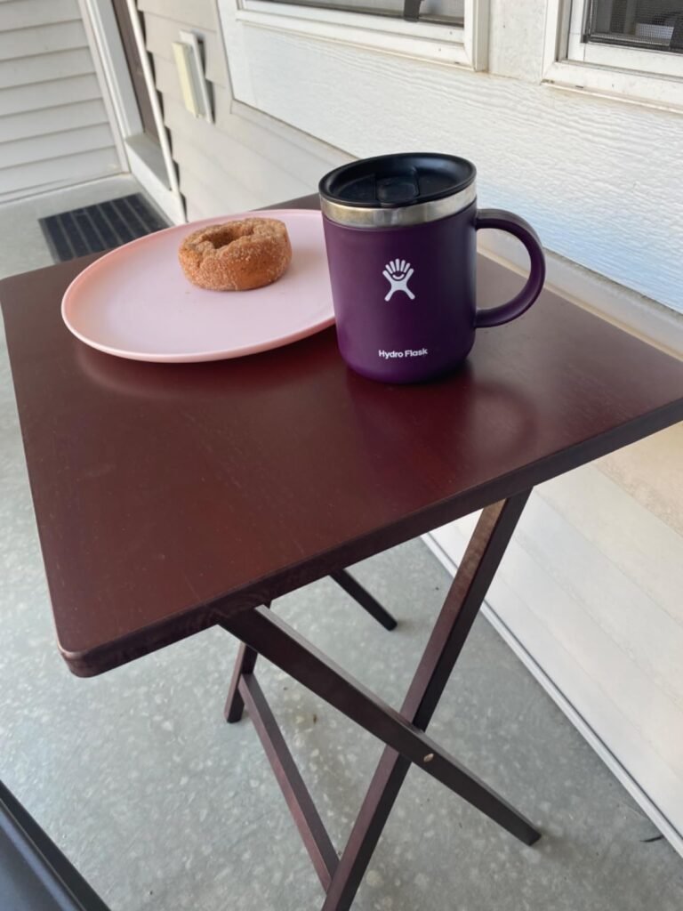 Mesa plegable de madera con una taza y un plato con un donut, colocada en una terraza.