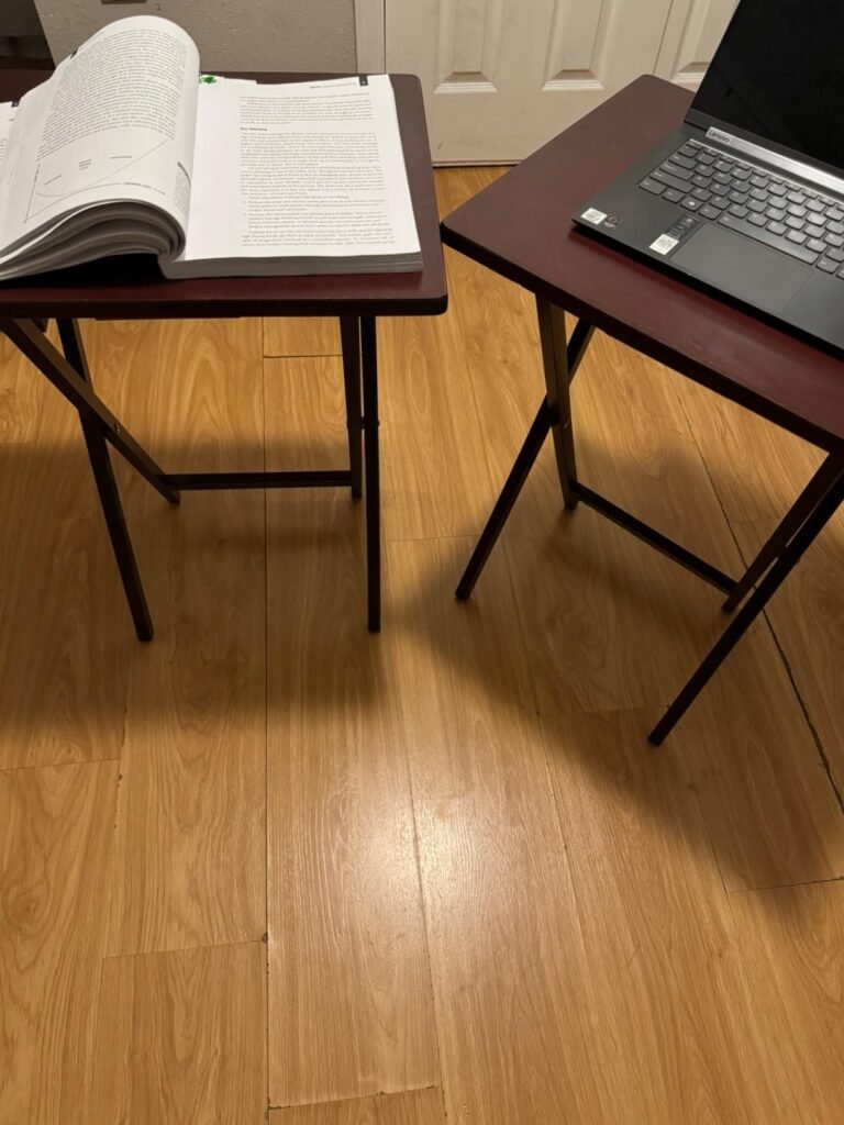 Dos mesas plegables de madera, una con un libro abierto y la otra con un portátil, sobre un suelo de madera clara.