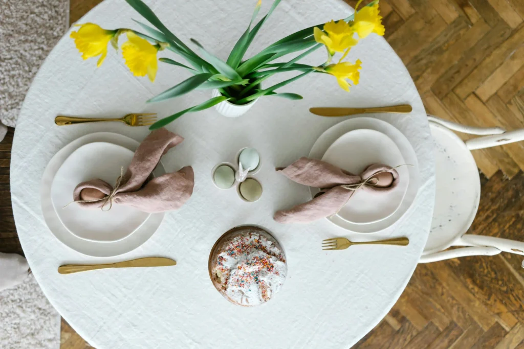 Mesa plegable redonda decorada con mantel blanco, platos de cerámica, servilletas de lino rosa, cubiertos dorados y un centro de mesa con flores amarillas, acompañada de un pastel cubierto de azúcar glas y confites.