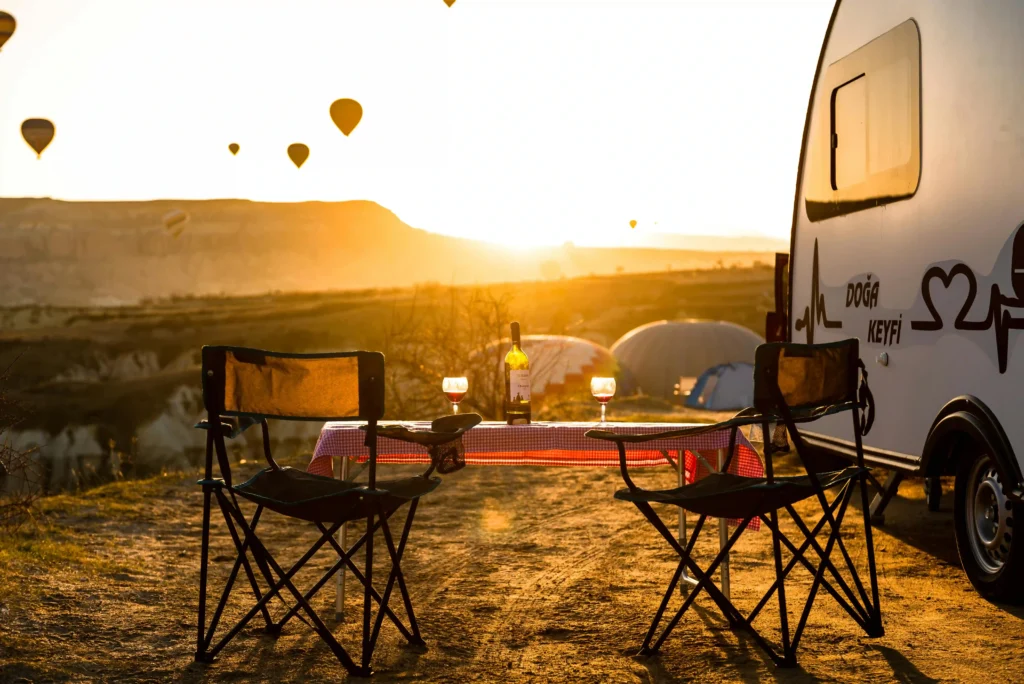 Escena de camping al atardecer con dos sillas plegables, una mesa con mantel, copas de vino y una botella, junto a una caravana y globos aerostáticos en el cielo.