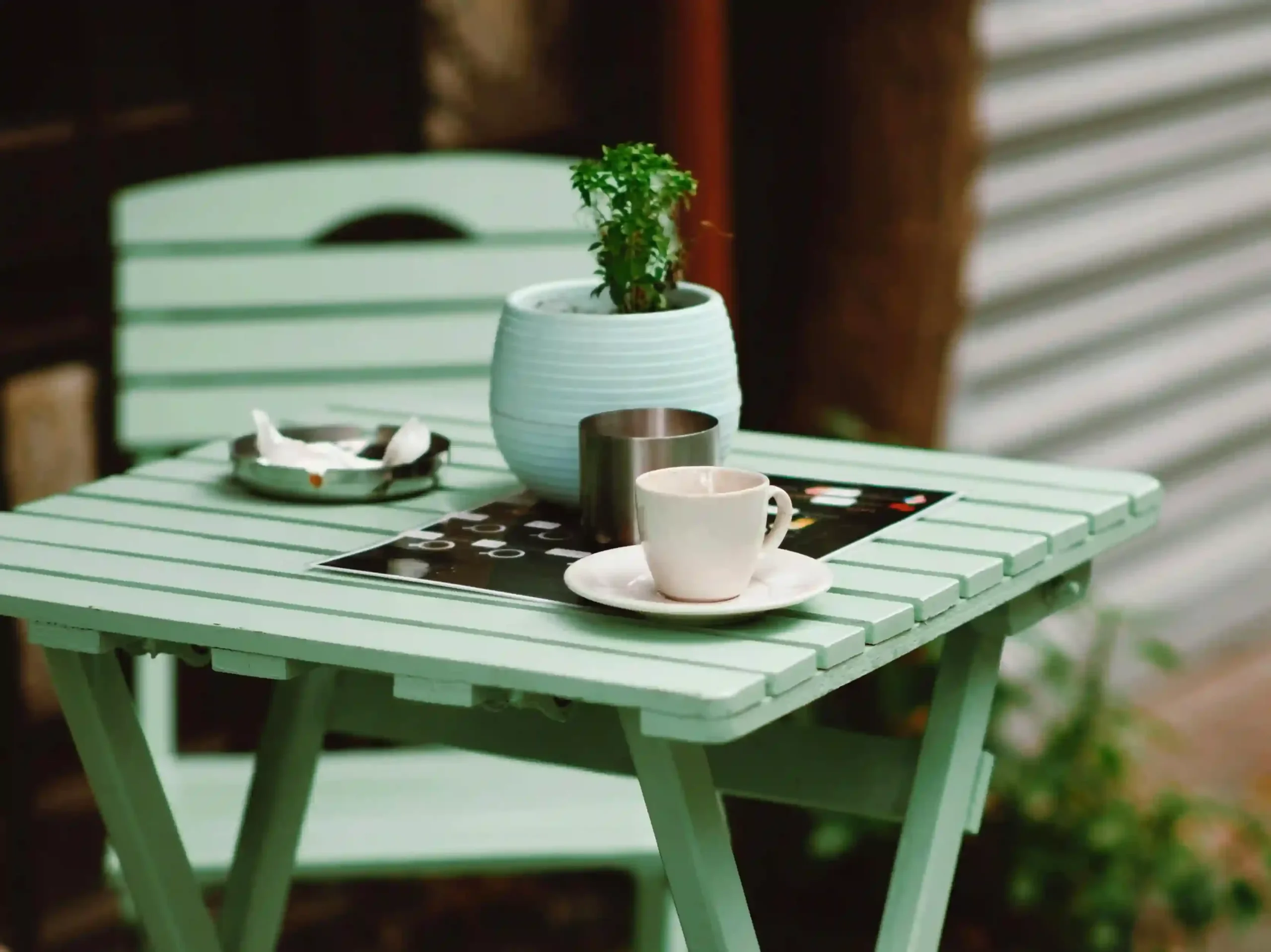 Mesa plegable de madera pintada de verde pastel, decorada con una taza de café, un pequeño macetero con planta y un cenicero, ubicada en un espacio al aire libre.