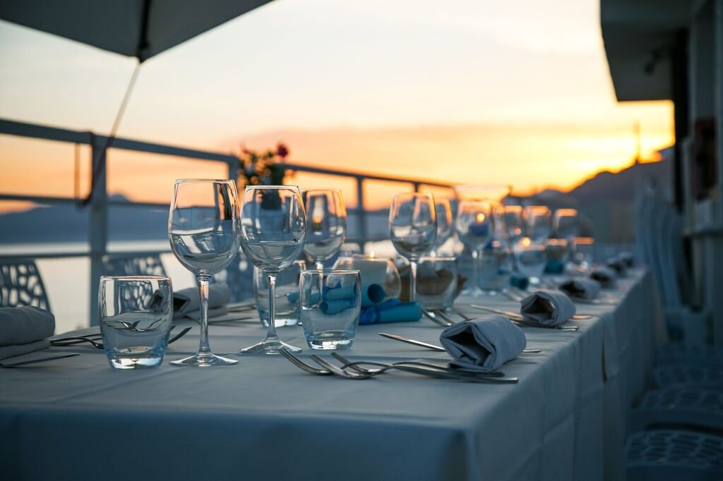 Mesa elegante al aire libre decorada con copas de cristal y servilletas, frente a un atardecer con vista al mar.