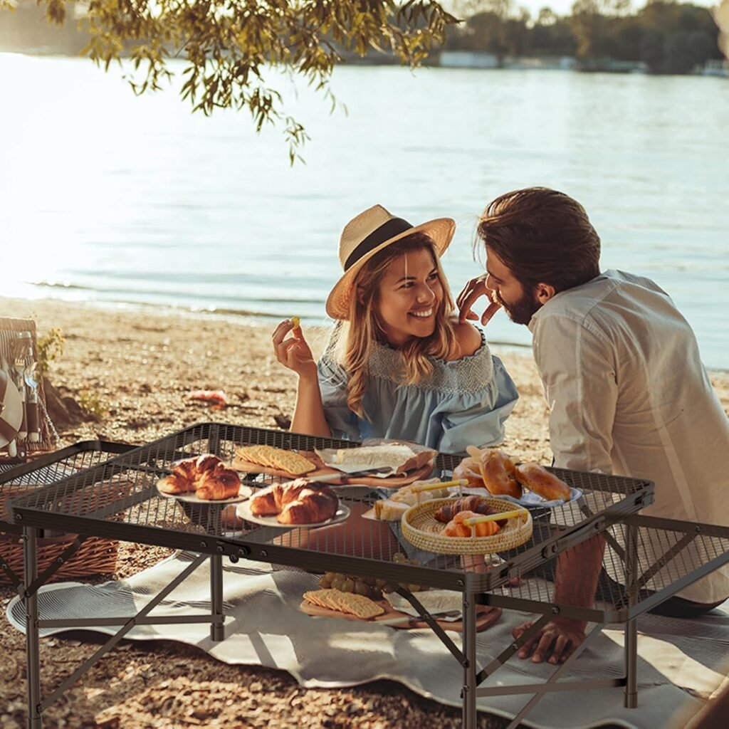 Pareja disfrutando de un picnic al aire libre junto a un lago con una mesa plegable de camping llena de comida.