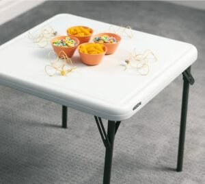 Mesa plegable Lifetime blanca con tazones de plástico naranja llenos de materiales para manualidades, como cuentas y macarrones, en un ambiente interior.