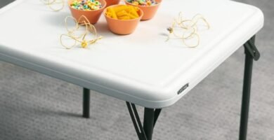 Mesa plegable Lifetime blanca con tazones de plástico naranja llenos de materiales para manualidades, como cuentas y macarrones, en un ambiente interior.