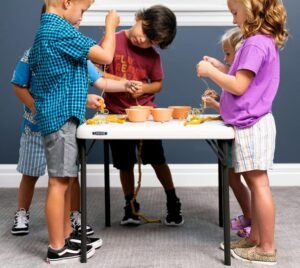 Niños pequeños participando en una actividad creativa con manualidades en una mesa blanca plegable en un ambiente interior.