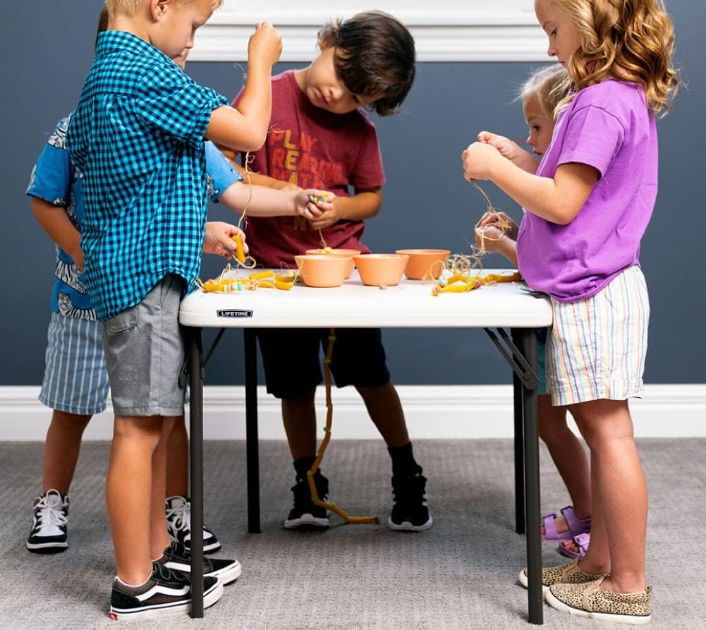 Niños pequeños participando en una actividad creativa con manualidades en una mesa blanca plegable en un ambiente interior.