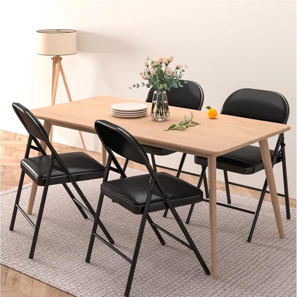 Comedor moderno con mesa de madera clara y cuatro sillas plegables negras, decorado con un florero con rosas, platos apilados y una lámpara de pie de trípode sobre una alfombra tejida.