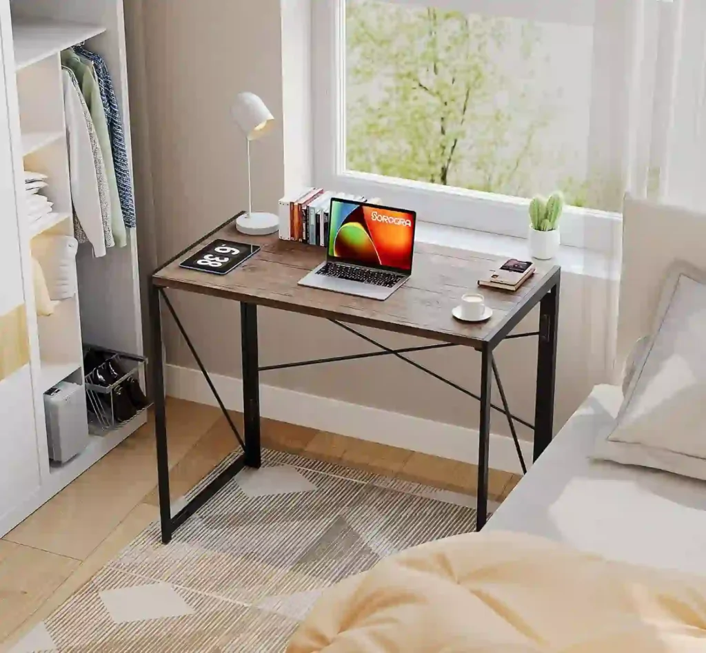 Mejor mesa plegable para escritorio. Construida de madera rústica con estructura metálica negra, ubicado junto a la ventana en un dormitorio, con laptop, libros, lámpara y taza.