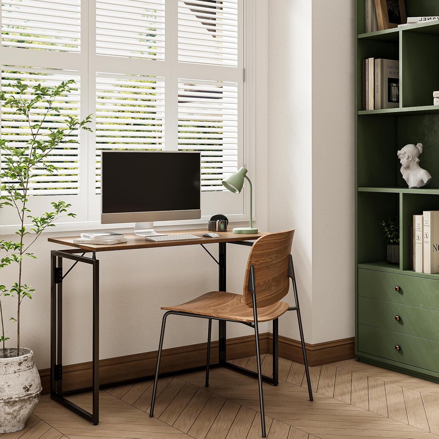Espacio de oficina en casa con escritorio plegable de madera y estructura metálica, silla de madera con patas negras, computadora de escritorio y lámpara verde, junto a una ventana con persianas blancas.