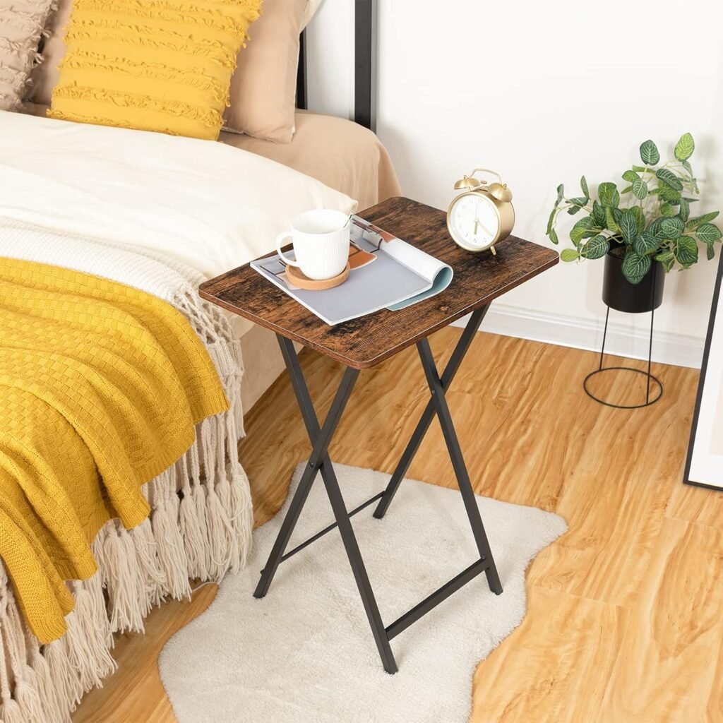 Mesa plegable pequeña auxiliar de madera rústica con patas metálicas negras junto a una cama con ropa de cama en tonos beige y amarillo, decorada con una taza, revistas y un reloj dorado.
