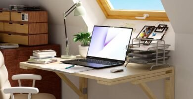 Escritorio flotante de madera clara montado en la pared bajo una ventana inclinada, con laptop, libros, lámpara de escritorio, organizador de documentos y silla giratoria blanca.