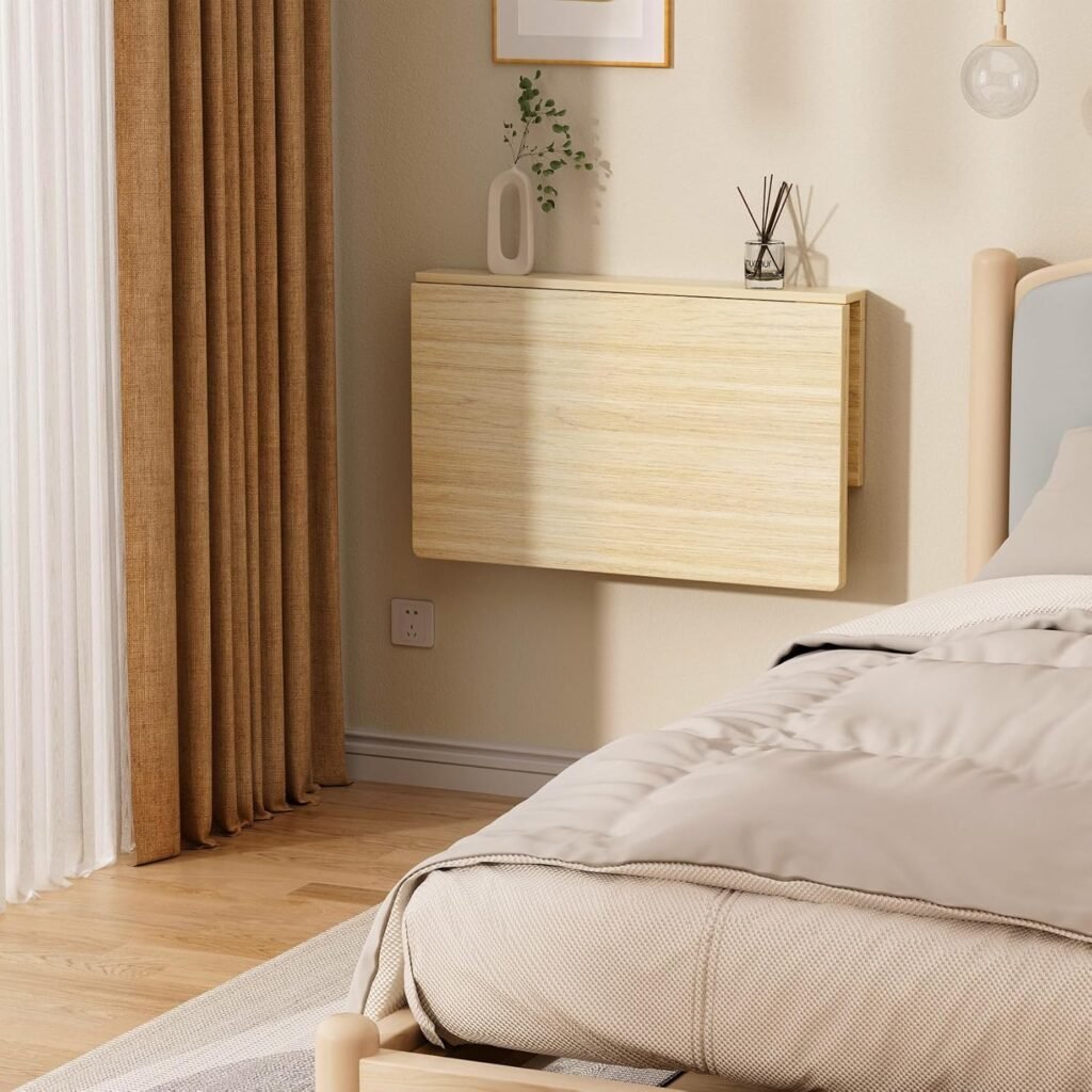 Escritorio plegable de madera clara montado en la pared de un dormitorio moderno, ubicada junto a la cama y decorada con un jarrón blanco y difusor de aroma.