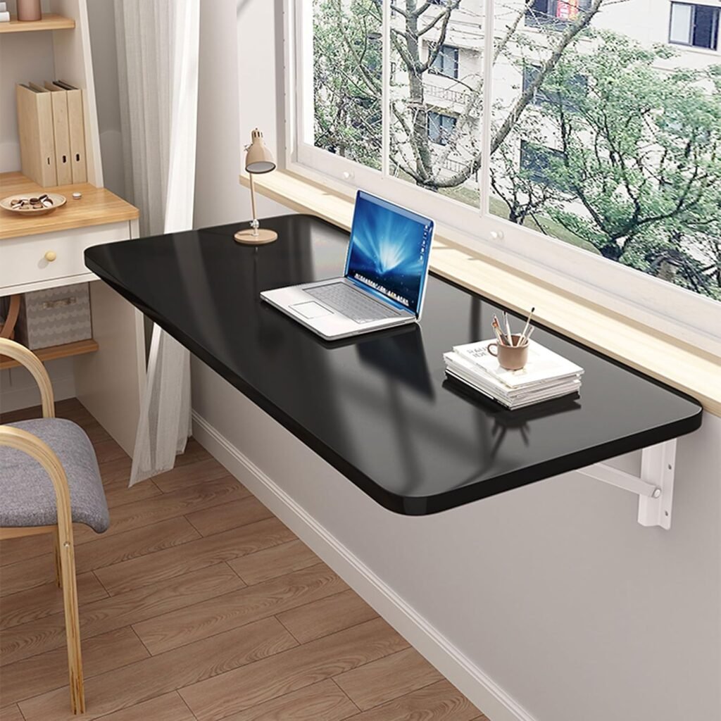 Escritorio plegable negro montado en la pared bajo una ventana, con una laptop, lámpara de escritorio, taza y cuadernos, en un espacio de trabajo moderno y luminoso.