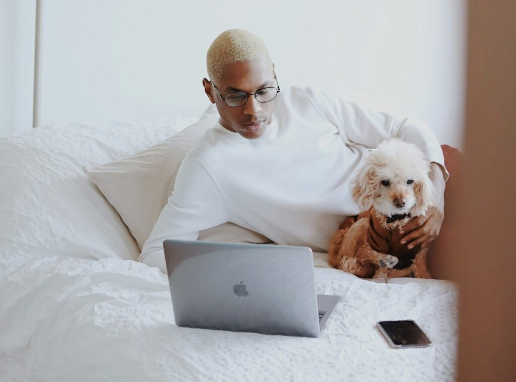 Hombre trabajando desde la cama con laptop y perro pequeño en un ambiente minimalista.