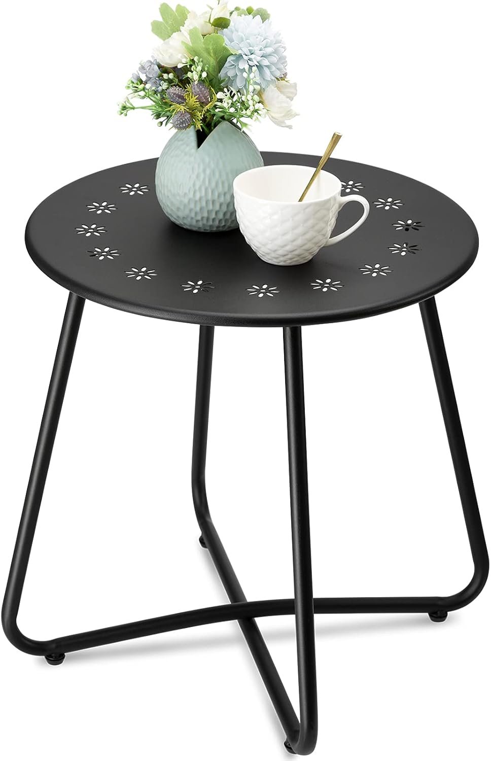 Mesa auxiliar redonda de metal negro con diseño de flores troqueladas en la superficie, decorada con una taza blanca y un florero azul con flores artificiales.