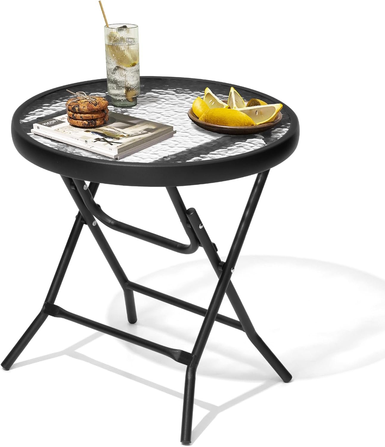 Mesa redonda plegable con estructura metálica negra y superficie de vidrio texturizado, decorada con una bebida, galletas, libros y un plato con rodajas de limón.