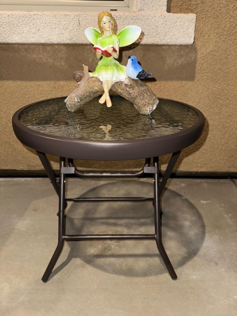 Mesa redonda plegable de vidrio con estructura metálica negra, decorada con una figura de hada sentada sobre un tronco junto a un pájaro azul, ubicada en un patio exterior.