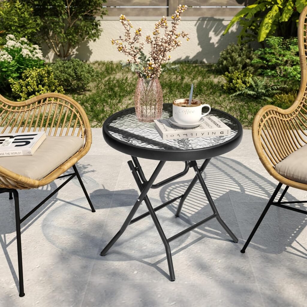 Mesa redonda plegable de vidrio con estructura metálica negra, colocada entre dos sillas de ratán con cojines, decorada con una taza de café, libros y un florero en una terraza con jardín al fondo.