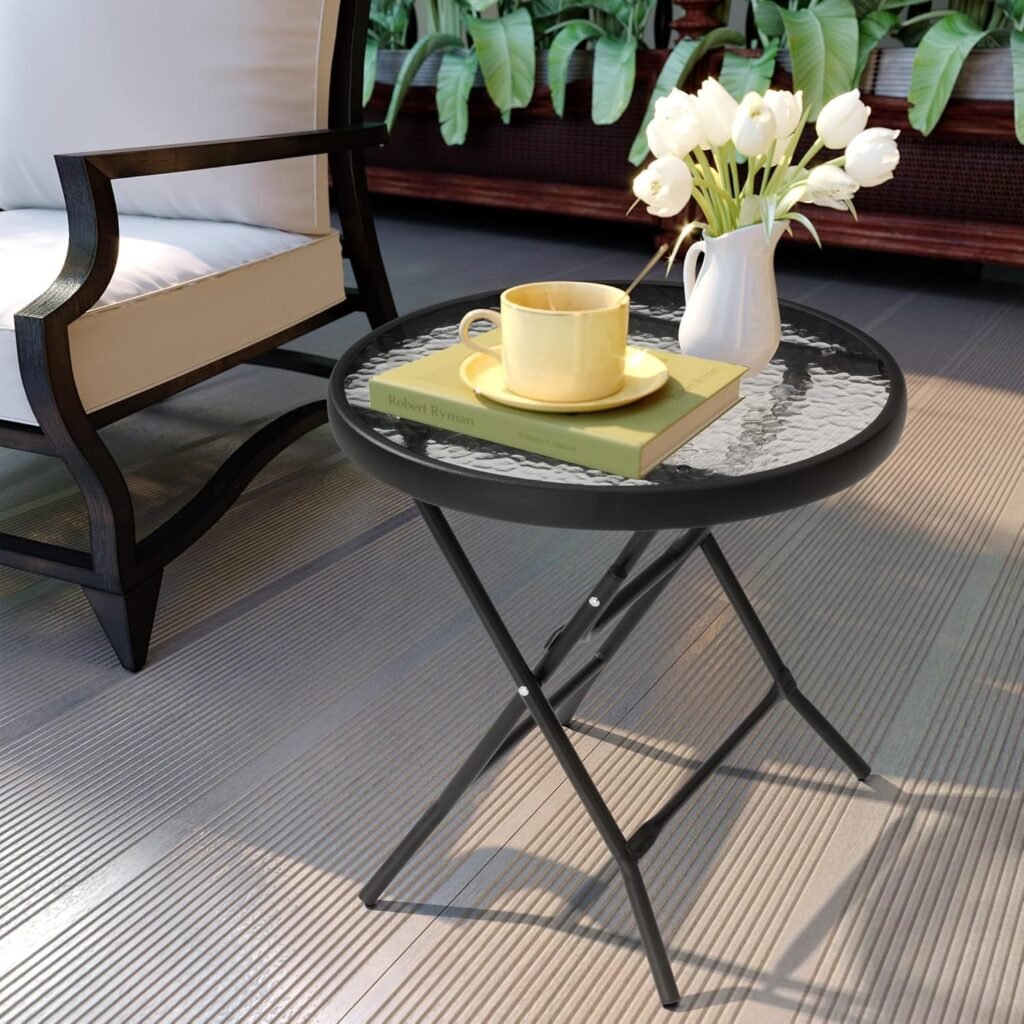 Mesa redonda plegable de vidrio con estructura metálica negra, decorada con una taza amarilla, libros y un florero con tulipanes blancos, junto a una silla de exterior acolchada.