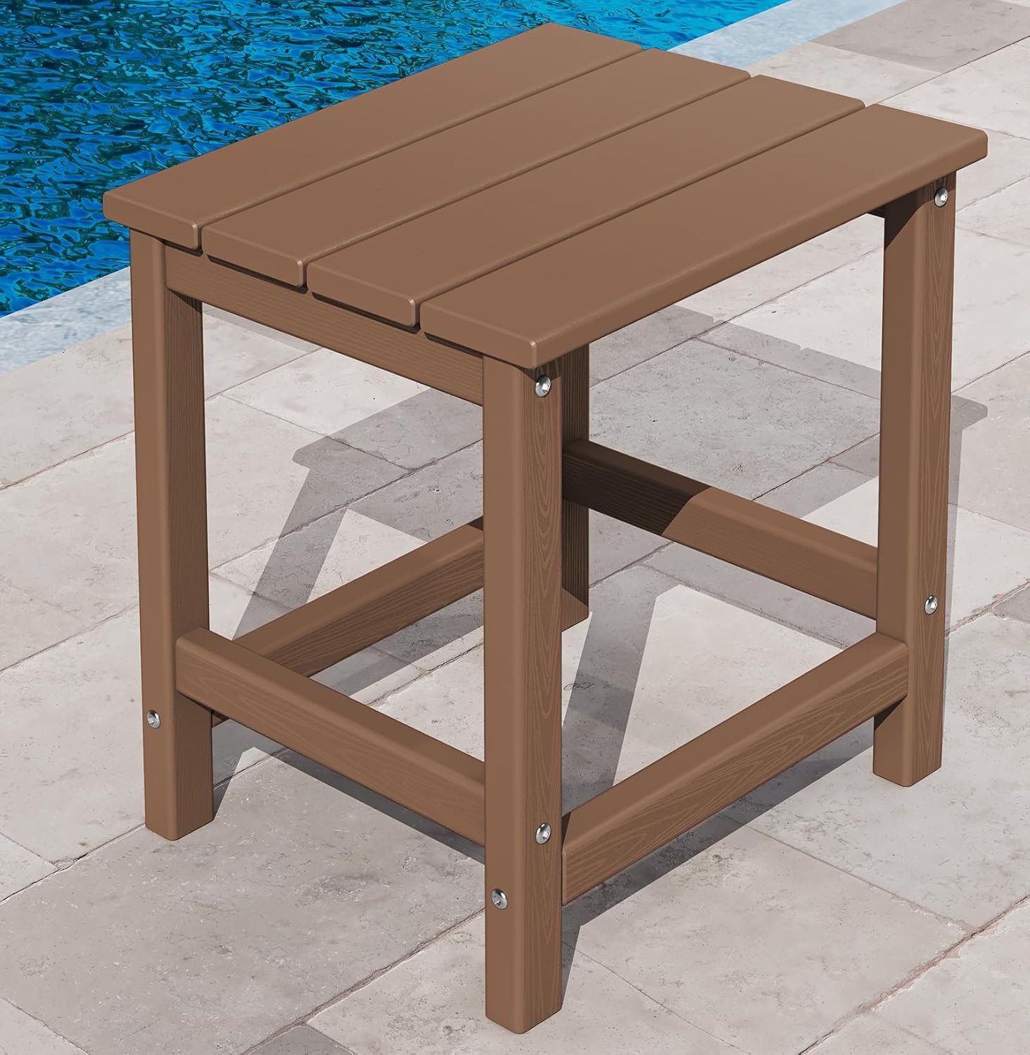 Mesa auxiliar cuadrada de exterior en color marrón, con diseño de listones en la cubierta y estructura robusta, colocada junto a una piscina sobre piso de piedra.