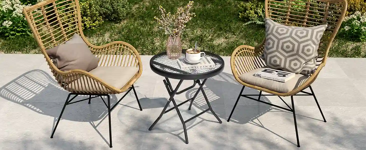 Conjunto de jardín con dos sillas de ratán con cojines y respaldo alto, acompañadas de una mesa redonda plegable negra con una taza de café, jarrón decorativo y libros, sobre una terraza con vegetación al fondo.