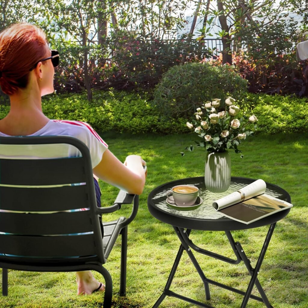 Mujer sentada en una silla de jardín contemplando la naturaleza, junto a una mesa redonda plegable de vidrio con estructura negra, decorada con una taza de café, revista enrollada y florero con rosas blancas.