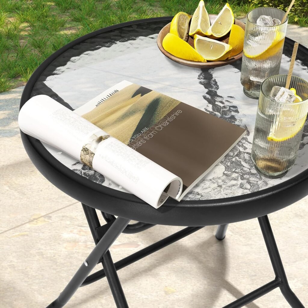 Mesa redonda plegable de vidrio con estructura metálica negra, sobre la que hay dos vasos con limonada, un plato con rodajas de limón y una revista enrollada, colocada en una terraza al aire libre.