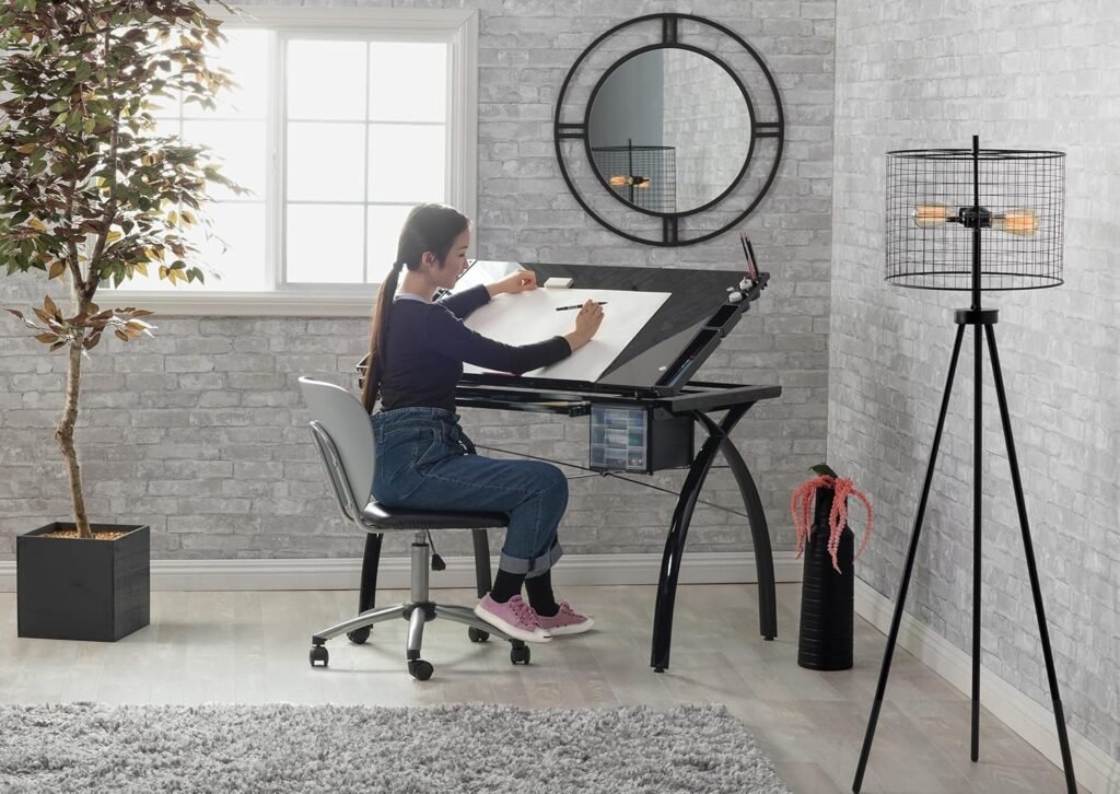 Mujer dibujando en un restirador de dibujo ajustable con estructura metálica negra y superficie inclinada, en una habitación moderna con pared de ladrillo gris, lámpara de trípode y espejo circular.