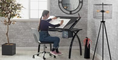 Mujer dibujando en un restirador de dibujo ajustable con estructura metálica negra y superficie inclinada, en una habitación moderna con pared de ladrillo gris, lámpara de trípode y espejo circular.